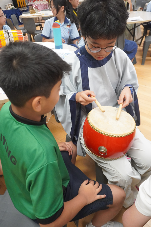 A student holding a small red drum while another child practices hitting it with wooden sticks.
